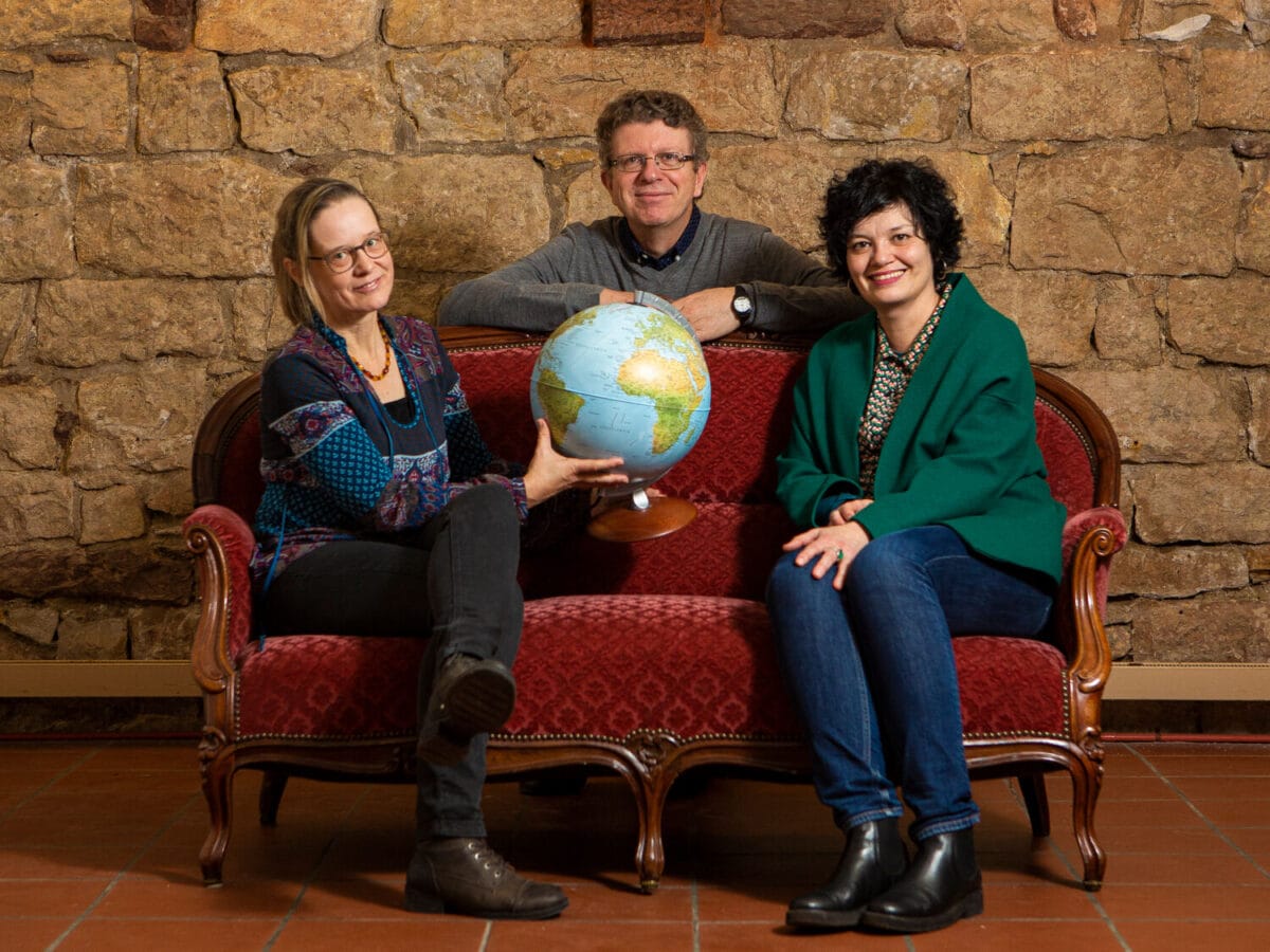 Alexandra Flury-Schölch and Claudia Buess with globe on a red sofa. Christian Weber looks over the backrest.