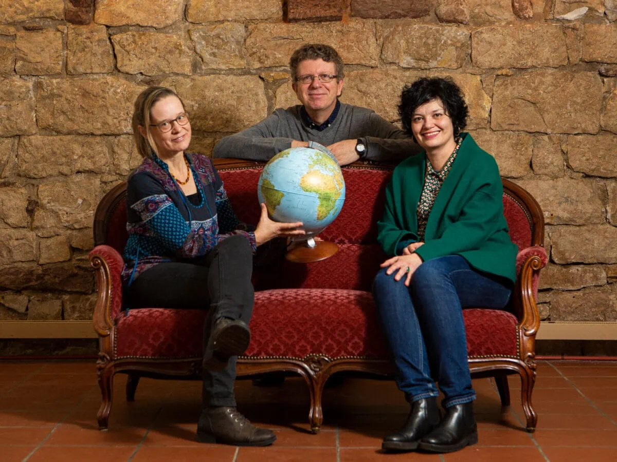 Alexandra Flury-Schölch et Claudia Buess avec un globe terrestre sur un canapé rouge. Christian Weber regarde par-dessus le dossier.